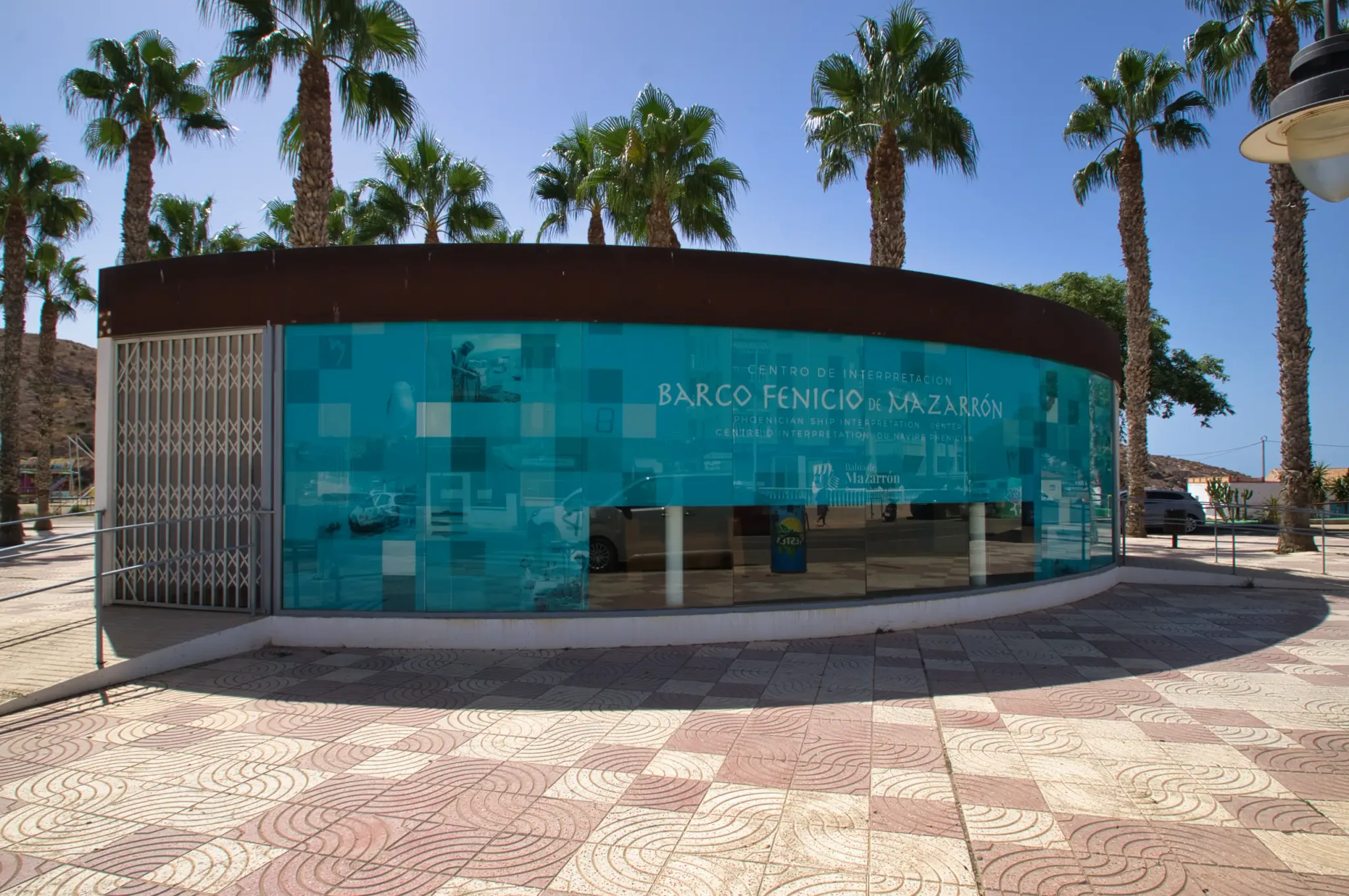 Curved turquoise exterior of the Phoenician Ship Interpretation Centre beneath palm trees in Puerto de Mazarrón