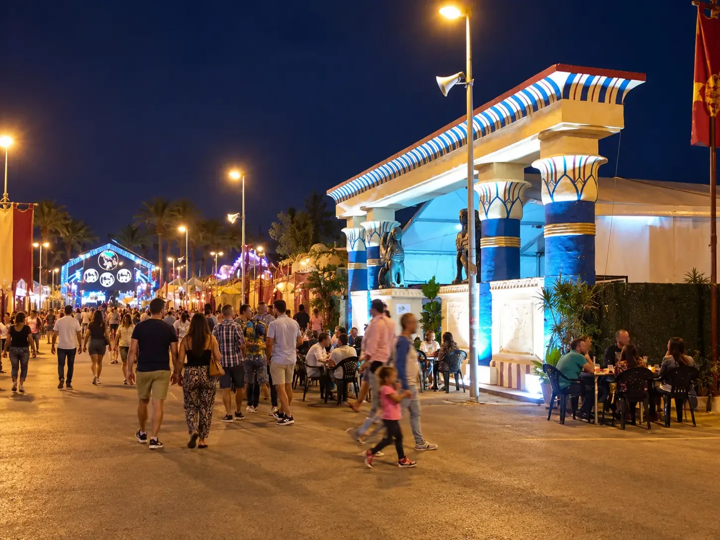 Visitors walking past an illuminated blue-and-white festival camp facade at night
