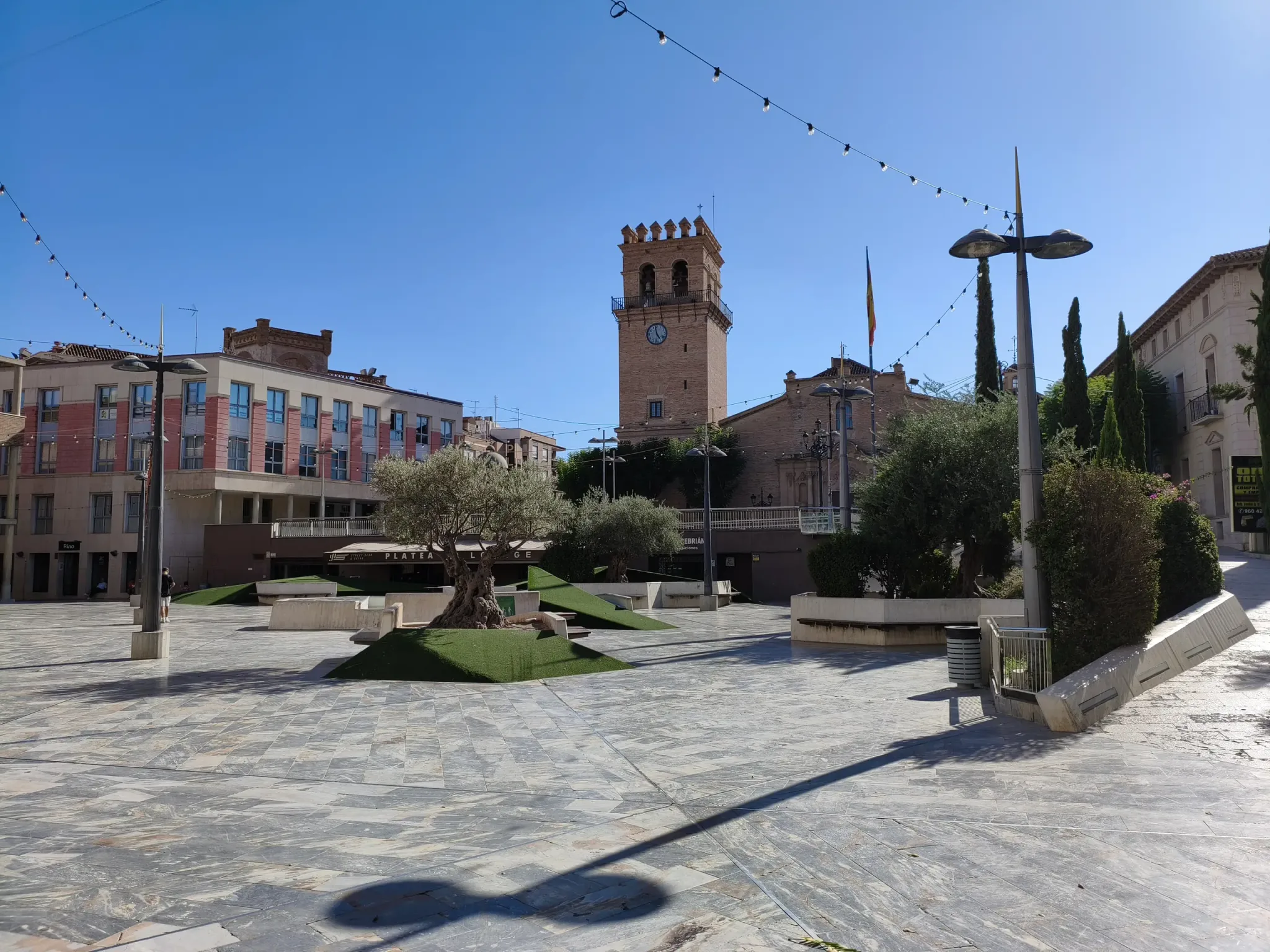 Place pavée ensoleillée avec des arbres et le clocher de l'église au centre de Totana